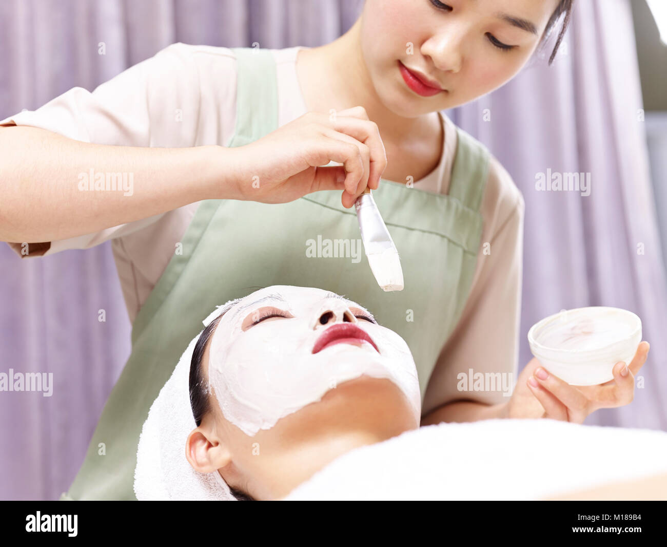 Asian estetista applicando bianco maschera per il viso con un pennello sul volto di una giovane donna. Foto Stock