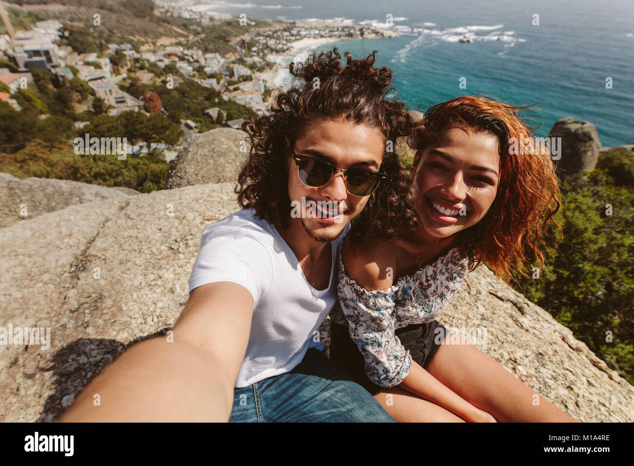 Giovane uomo e donna seduta insieme sulla cima della scogliera prendendo self portrait. Coppia felice seduto sulla cima della montagna e prendendo selfie. Foto Stock