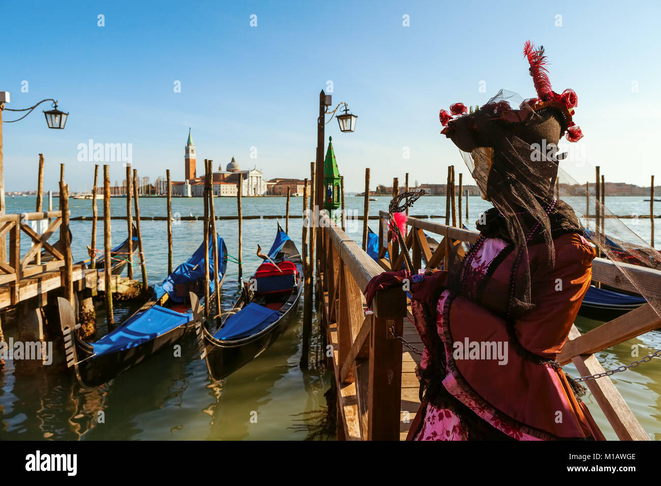 Il partecipante non identificato vestito in costume si erge nei pressi di gondole sul Canal Grande come chiesa di San Giorgio Maggiore sullo sfondo di Venezia. Foto Stock