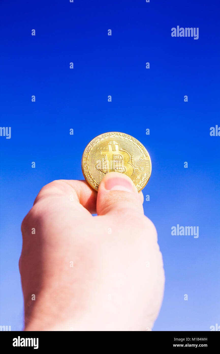 Crypto valuta. Bitcoin. Una mano sollevata detiene una moneta d'oro bit moneta contro il cielo blu. I raggi del sole si riflettono nella stessa moneta. Foto Stock