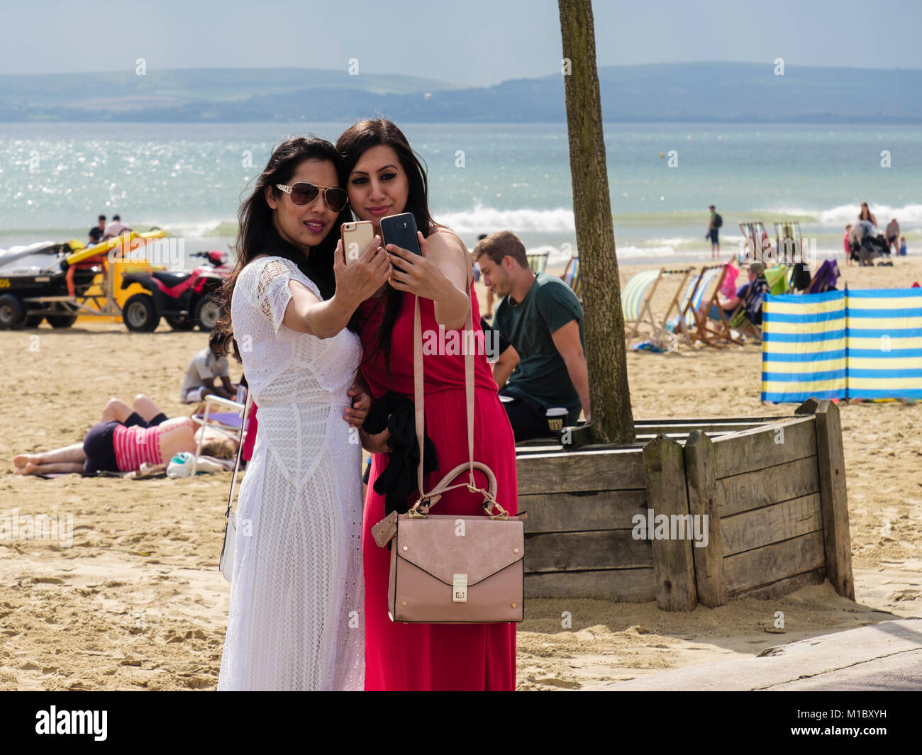 Due ben vestiti giovani donne millenario tenendo selfies insieme utilizzando smartphone su una spiaggia in tarda estate. Bournemouth Dorset England Regno Unito Gran Bretagna Foto Stock