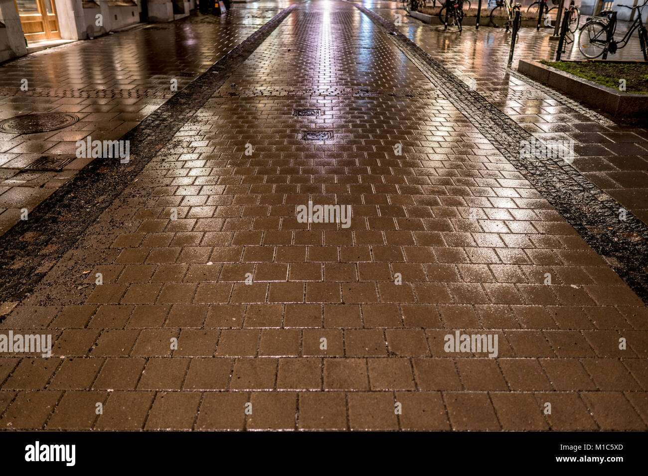Notte piovosa su una strada pedonale con pavimentazione in ciottoli di luci e dalla riflessione di edifici vicini e illuminazione della strada. Biciclette perked. Foto Stock
