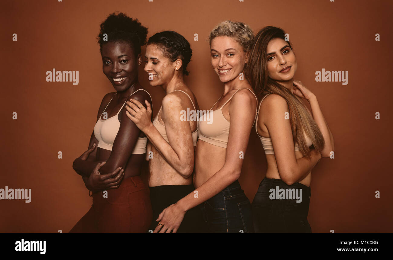 Modelli femmina con skin differenti in piedi insieme e sorridente. Gruppo di felice giovani donne su sfondo marrone. Foto Stock