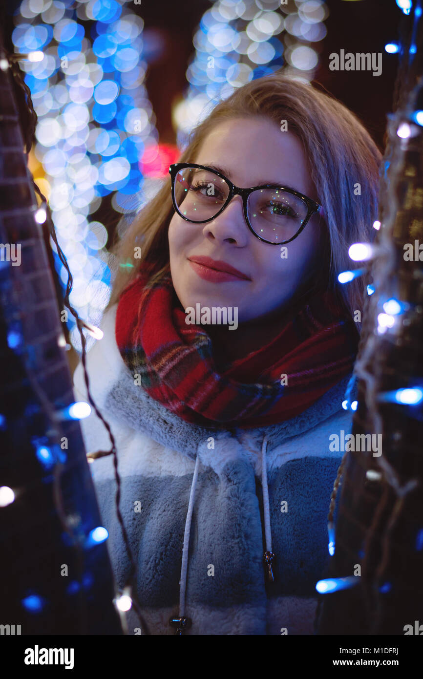 Una giovane ragazza attraente in bicchieri e una calda sciarpa passeggiate in città fiera. Sorride. Ritratto di notte tra ghirlande. Foto Stock