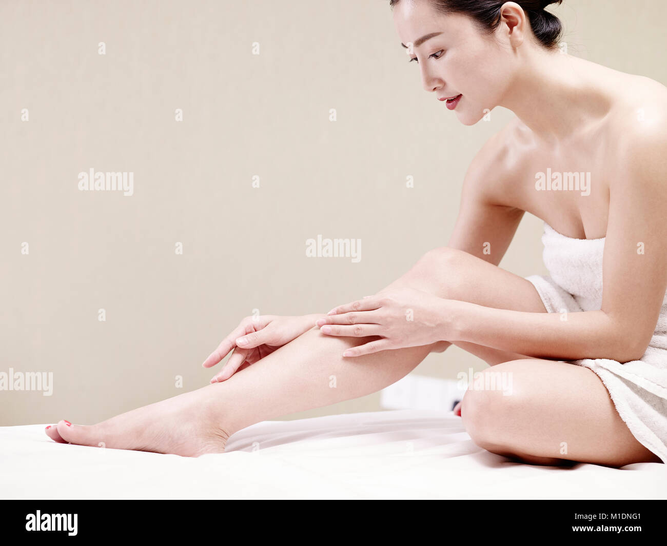 Bella giovane donna asiatica con una pelle morbida avvolto in asciugamano bianco seduta sul letto di applicare la lozione per il corpo. Foto Stock