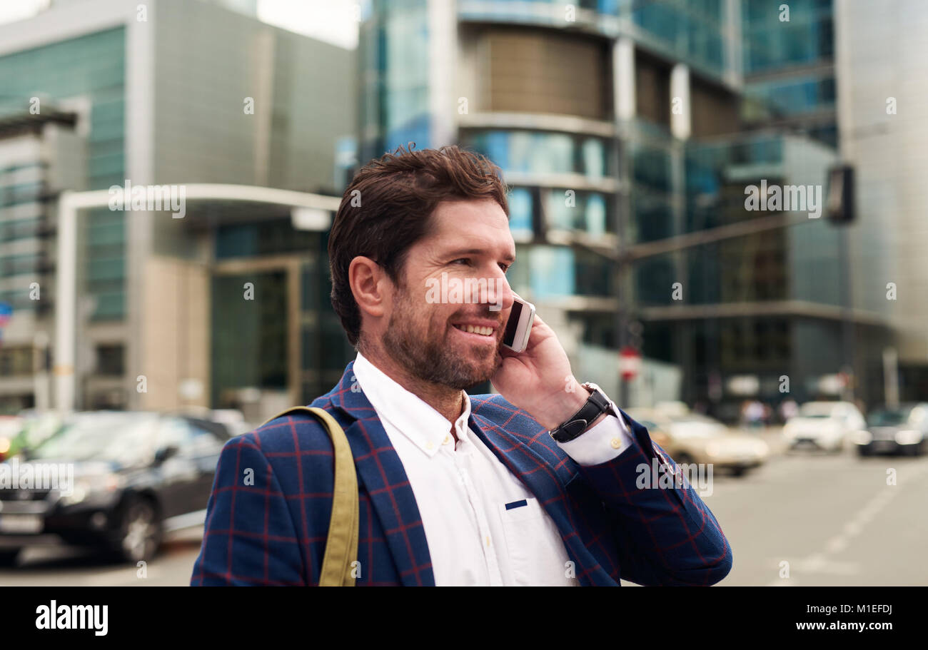 Imprenditore sorridente a piedi nella città di parlare sul suo cellulare Foto Stock