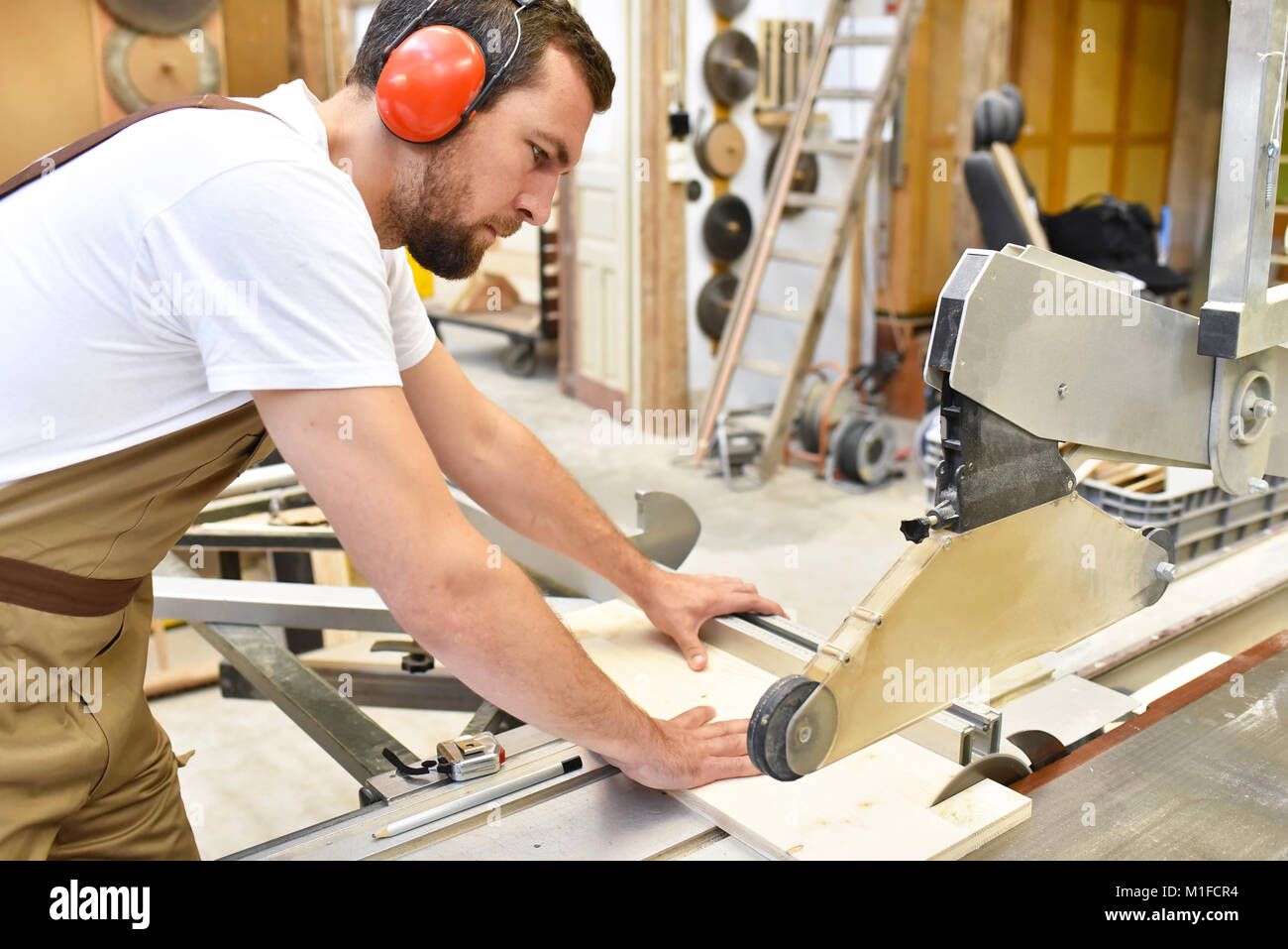 Gentile falegname con protezioni per le orecchie e gli indumenti da lavoro lavorando su una sega in officina Foto Stock