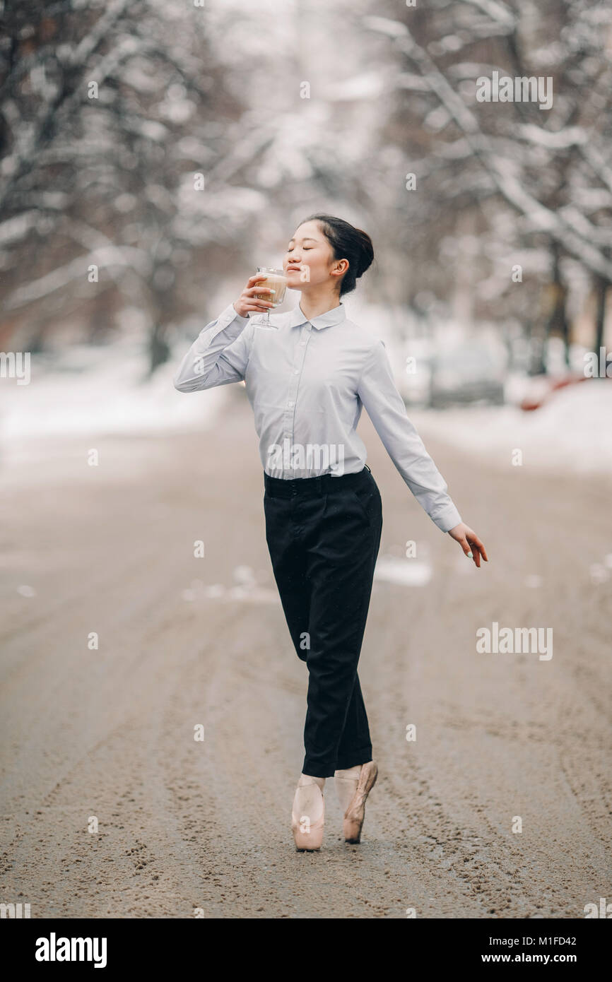 Ballerina vestita di nero i pantaloni e camicia bianca con un bicchiere di  caffè caldo raf nelle sue mani è in piedi accanto alla strada di città  contro la neve e inverno