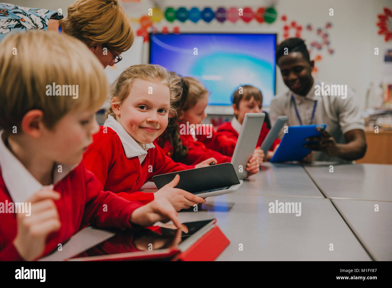 I bambini della scuola elementare sono in ckassroom, apprendimento e utilizzo di digital compresse. Una delle ragazze è sorridente alla fotocamera. Foto Stock