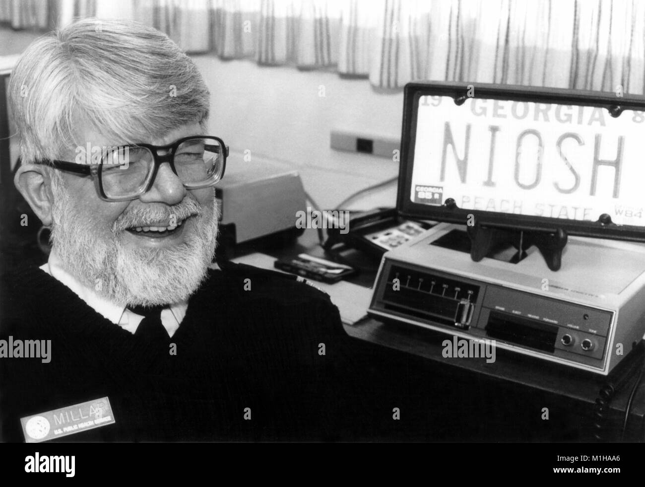 Headshot informale di J. Donald Millar, M.D. un epidemiologo e direttore dell'Istituto nazionale della salute e della sicurezza sul lavoro, ridendo accanto a un NIOSH targa, 1985. Immagine cortesia CDC. () Foto Stock