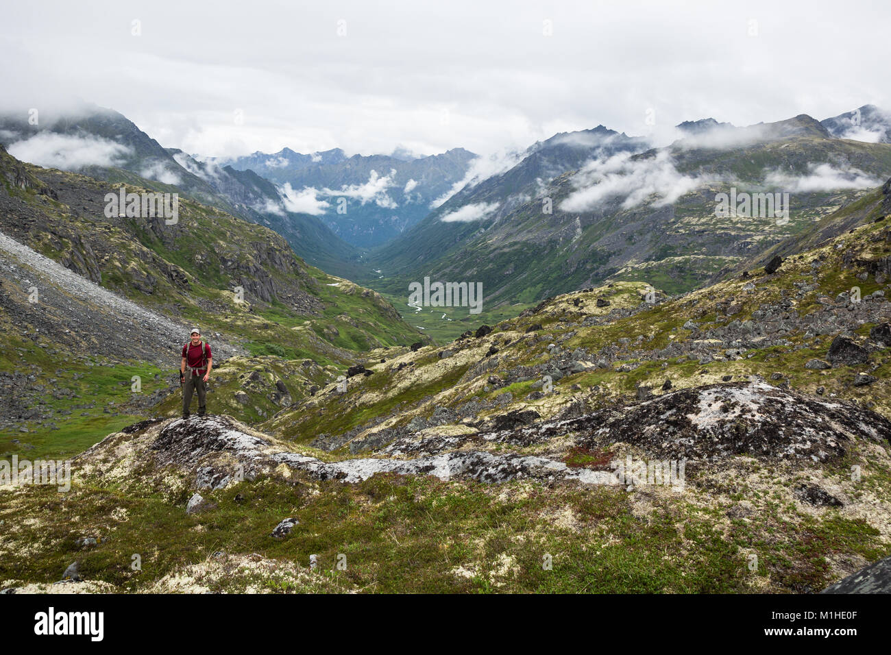 Un giovane maschio sta con il suo zaino e bastoni per escursioni su un punto di vista rocciosa che si affaccia su un fiume glaciale di valle in Hatcher Pass area dell Alaska Foto Stock