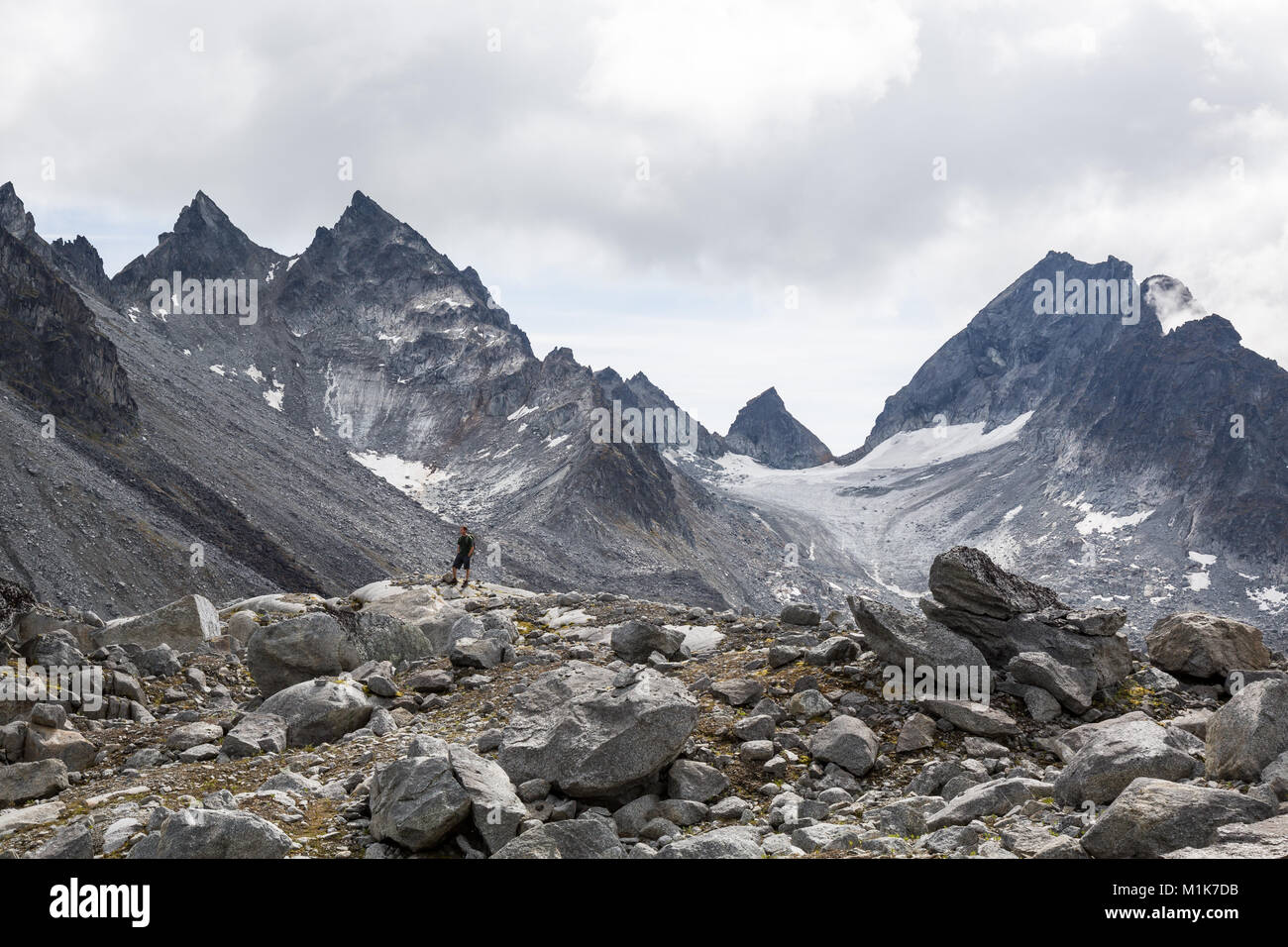 Un escursionista con le mani in tasca si erge su una collina rocciosa al di sotto di un panorama alpino di picchi rocciosi, Glacier Ice e ghiaioni piste Foto Stock