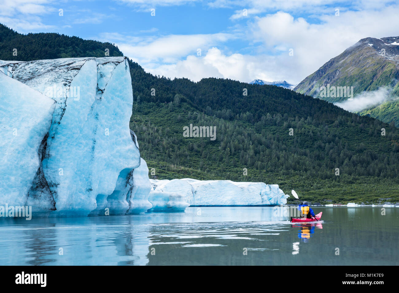 Un uomo in rosso le pale in kayak attraverso perfettamente ancora acqua del ghiacciaio Spencer terminale del lago. Il ghiacciaio di torri di ghiaccio overhead e iceberg può Foto Stock