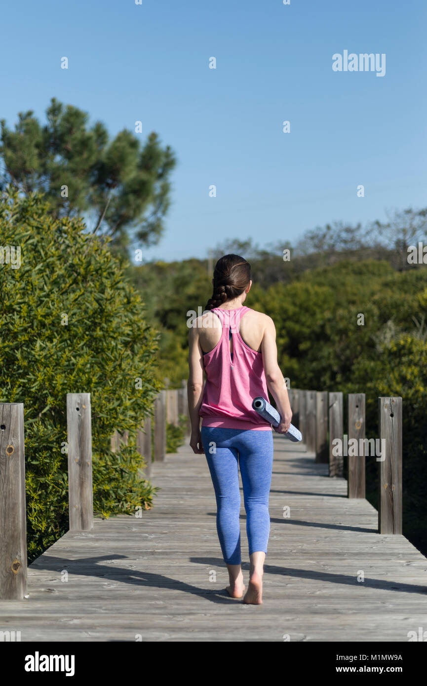 La donna che porta un arrotolato materassino yoga, esterno su un ponte di legno. Vista posteriore Foto Stock