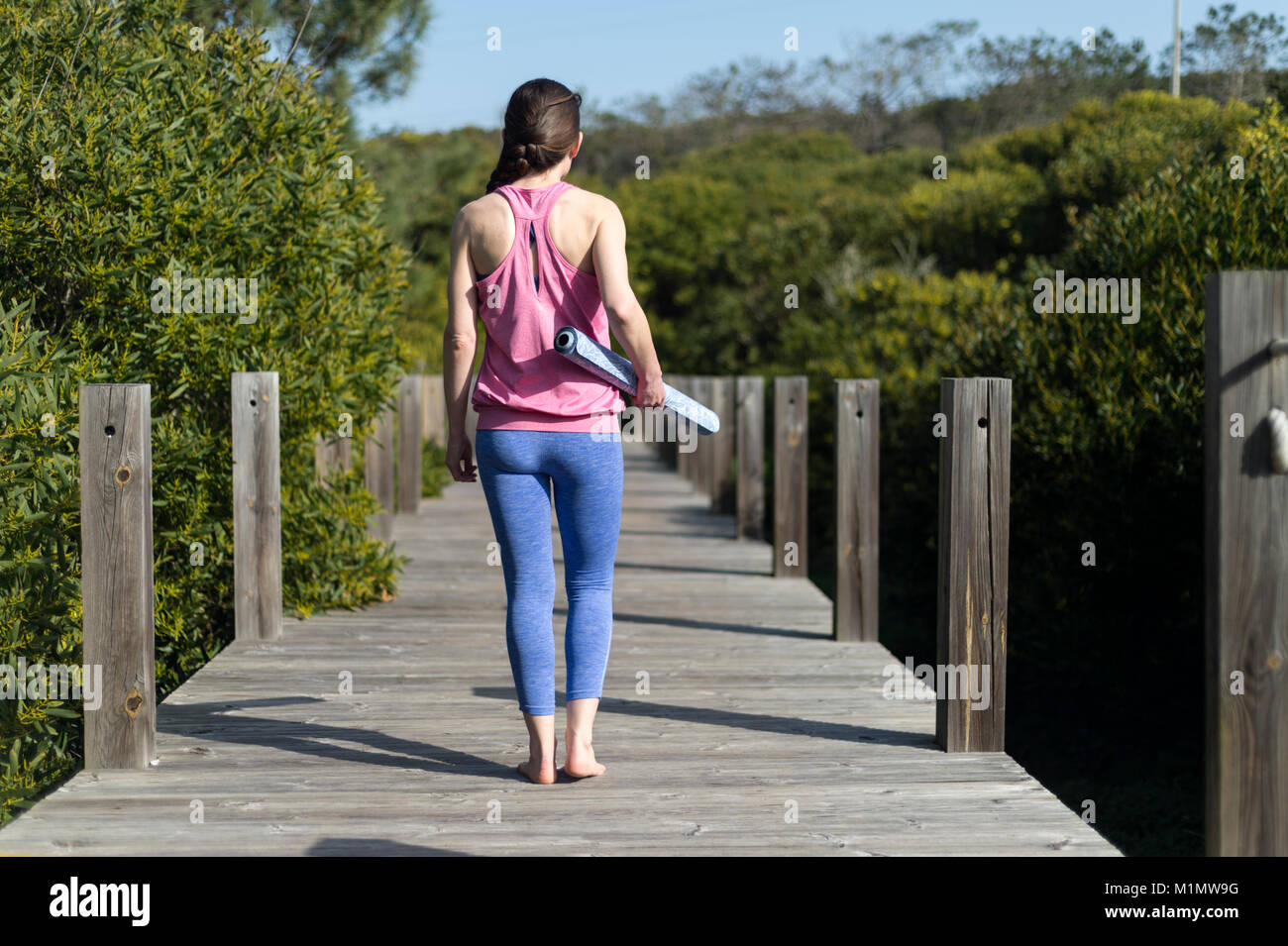 La donna che porta un arrotolato materassino yoga, esterno su un ponte di legno. Vista posteriore Foto Stock