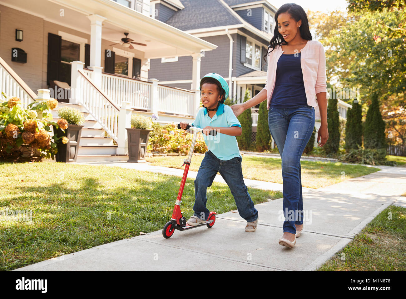 La madre cammina con Figlio come lui corre in scooter lungo il marciapiede Foto Stock