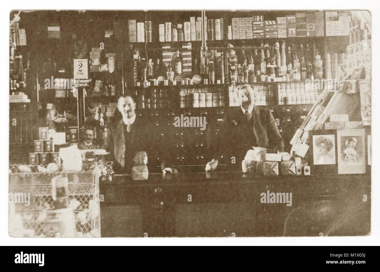 Originale cartolina dei primi del 1900 di 3 assistenti maschili del negozio, con baffi, all'interno di un negozio dietro il bancone, molte vecchie marche di prodotti, , Regno Unito circa 1912. Foto Stock