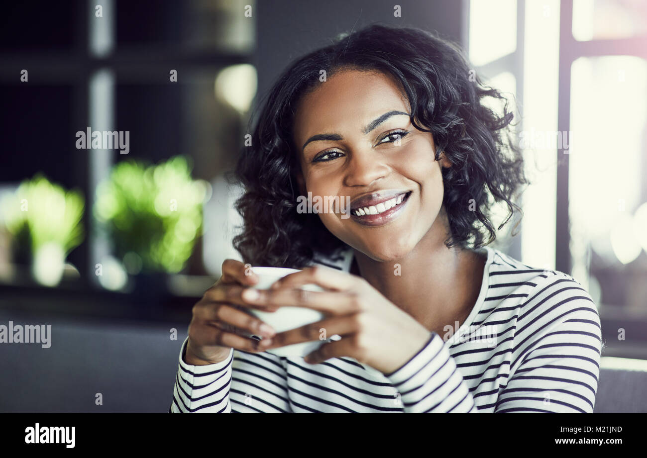 Giovane donna africana sorridente mentre è seduto da solo a un tavolo in un bar godendo di una nuova tazza di caffè Foto Stock