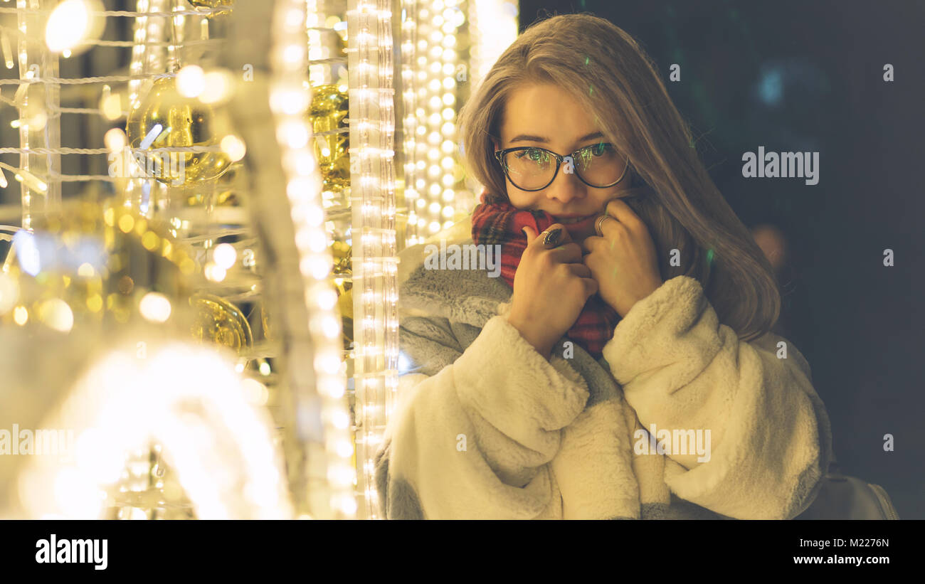 Un giovane parigamba dolce ragazza con gli occhiali e una calda sciarpa da passeggiate al centro commerciale su una vacanza. Sorride e raddrizza la sciarpa. Ritratto di notte accanto Foto Stock