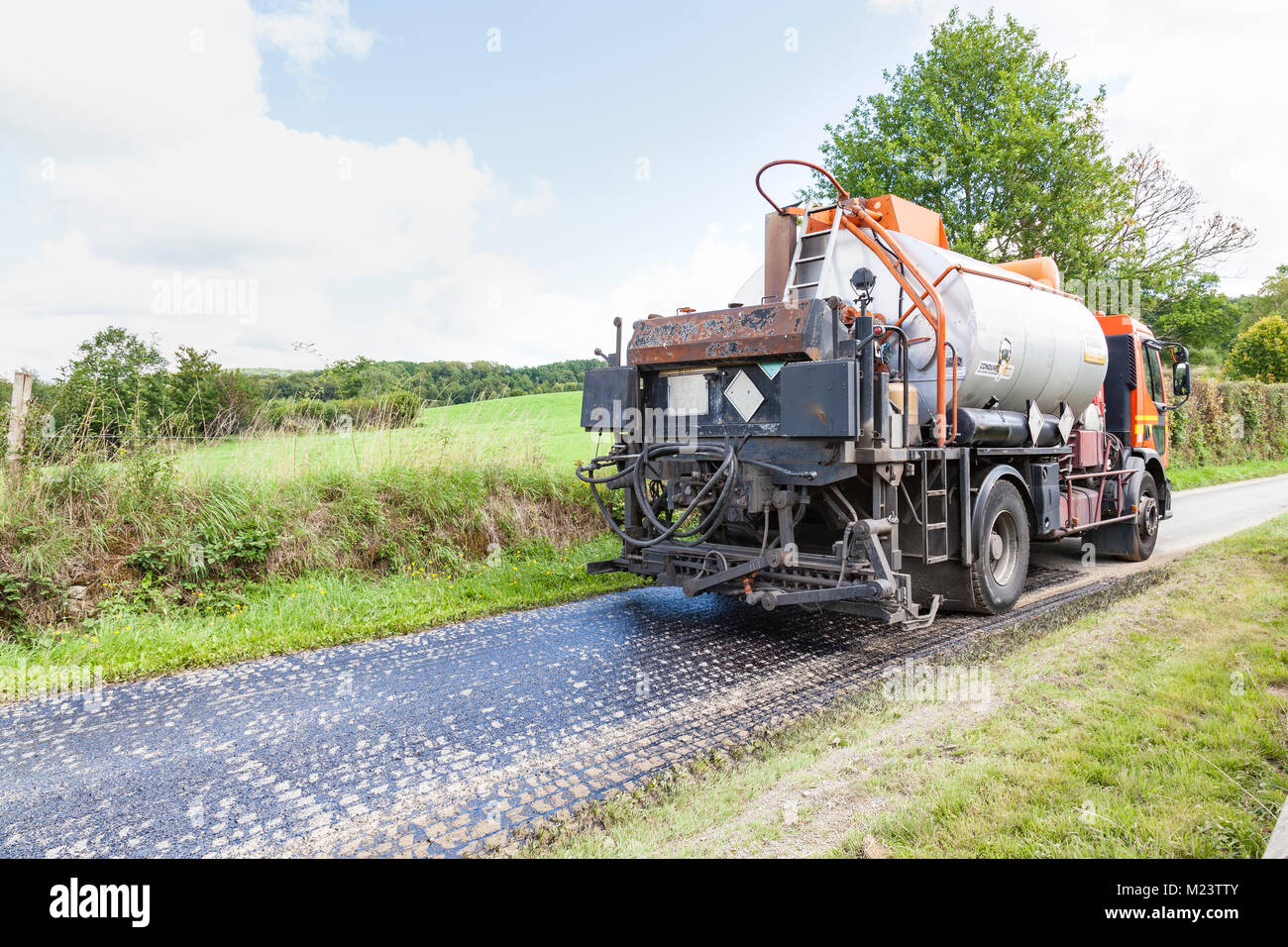Tanker affiorare una strada rurale con catrame liquido per formare una base di aderenza della premiscela asfalto che viene arrotolato e compattato sulla parte superiore durante la manutenzione Foto Stock