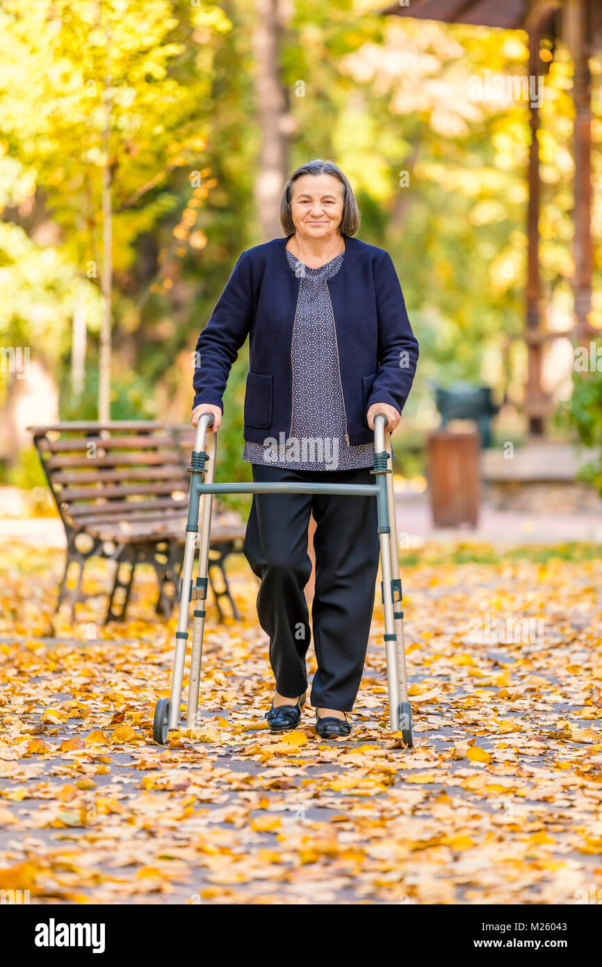 Senior donna passeggiate all'aperto con walker in autunno park e sorridente alla fotocamera. Foto Stock