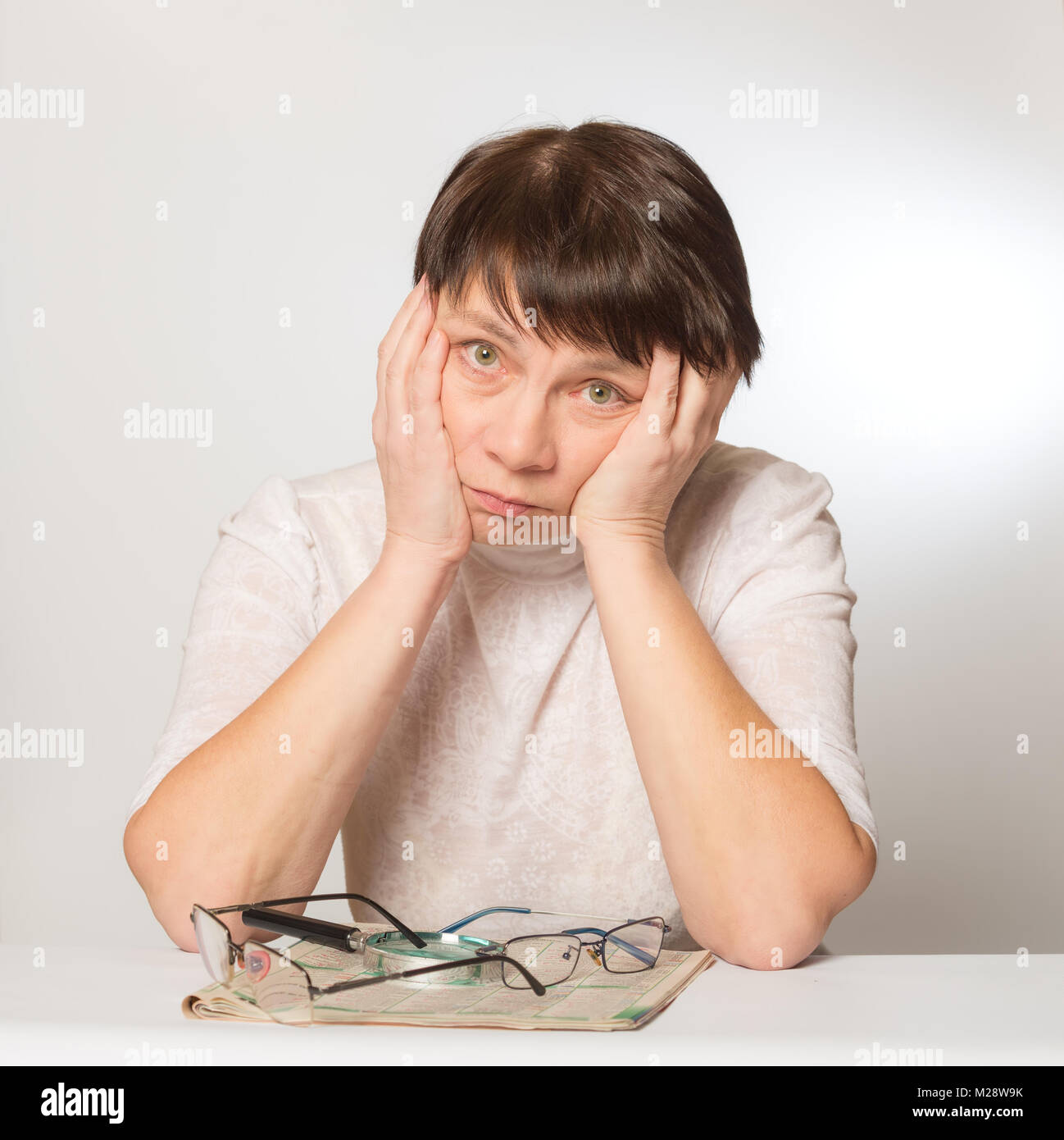 Frustrati donna mantiene la sua testa nelle sue mani. Sul tavolo di fronte a lei è un giornale, due bicchieri e una lente di ingrandimento. Scarsa visione, occhiali ar Foto Stock