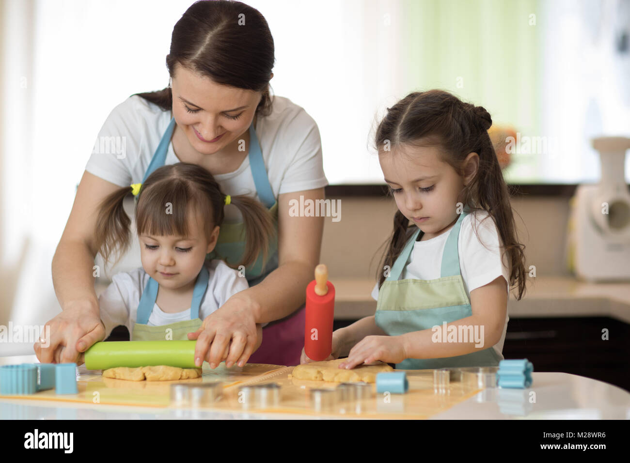 Bambini cute ragazze e le loro belle mom in grembiuli sono la riproduzione e ridere mentre impastare la pasta in cucina Foto Stock