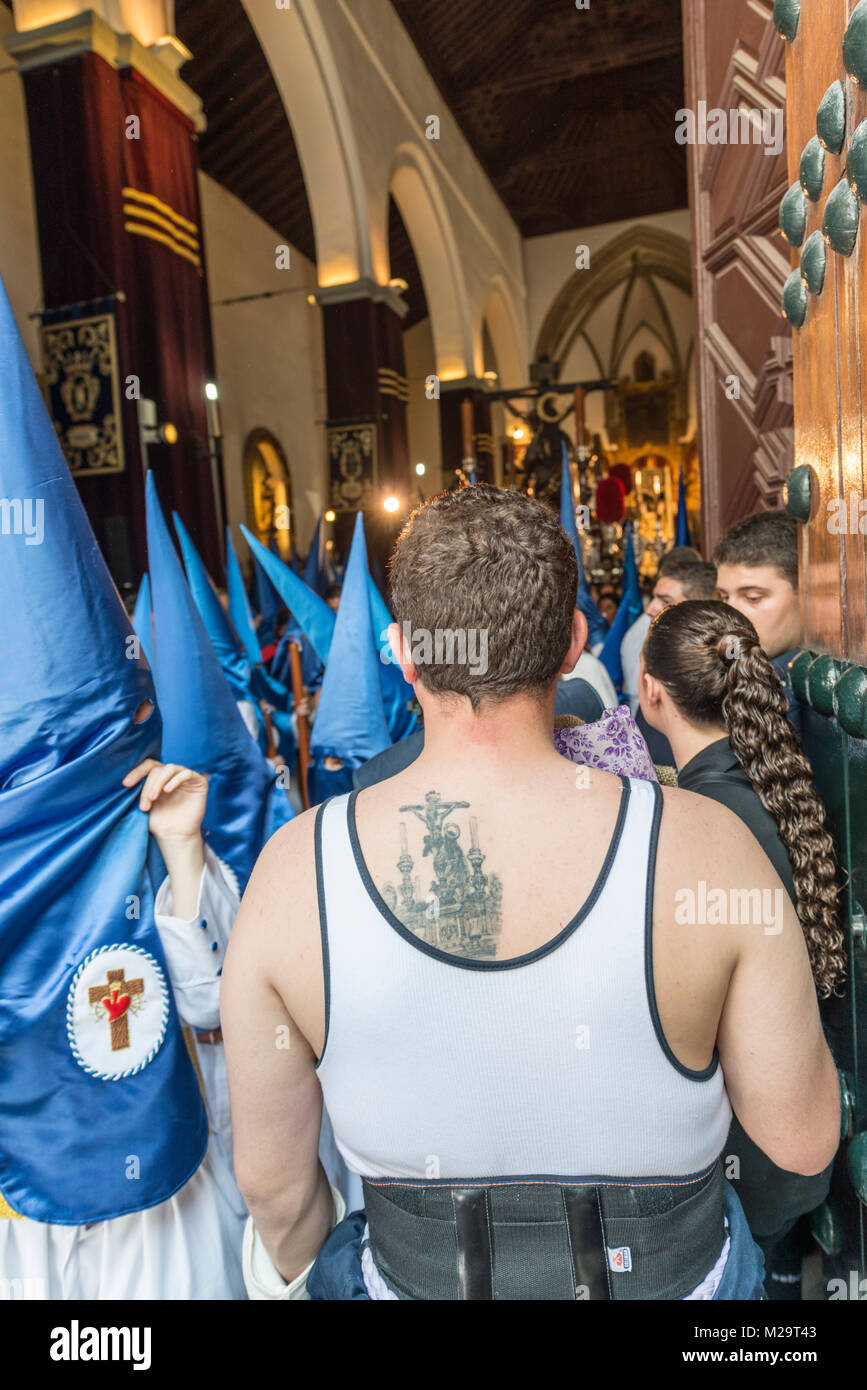 Portatore di fratellanza di 'La Hiniesta' con un tatuaggio sul suo retro osservata dalla porta della chiesa la partenza del galleggiante del Cristo del suo Foto Stock