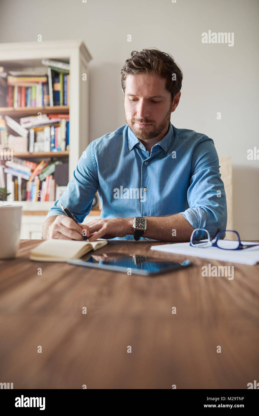 Giovane uomo seduto da solo a un tavolo nel suo soggiorno a scrivere le idee in un blocco note mentre si lavora da casa Foto Stock