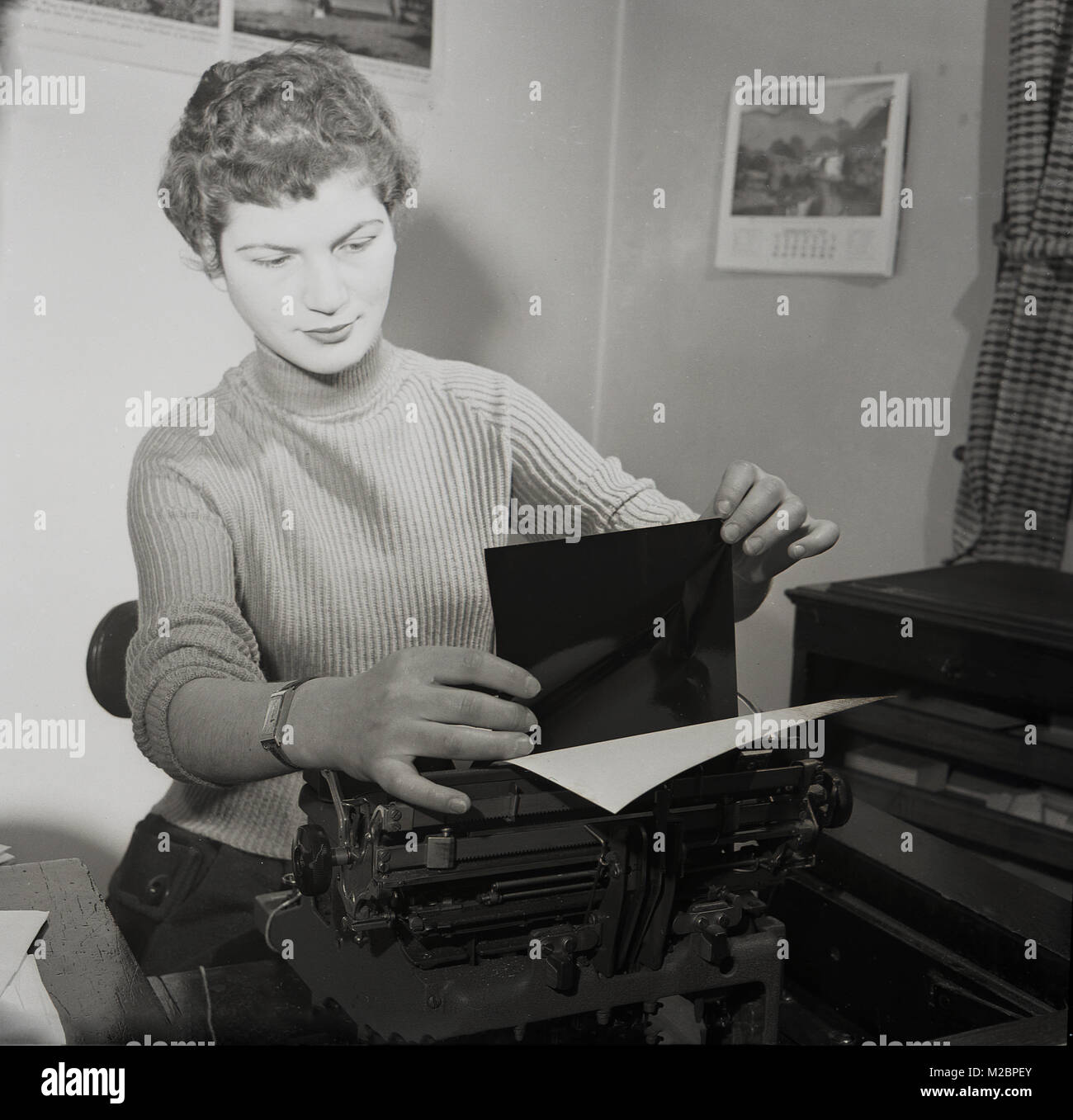 Degli anni Cinquanta, foto storiche di una giovane donna seduta a una scrivania in ufficio un carbonio di alimentazione carta in un manuale dattiloscritto, al fine di ottenere una copia o una impressione dalla lettera ella sarà tyoing fino, Inghilterra, Regno Unito. Nei giorni prima di dati elettronici come l'e-mail, alle aziende di inviare lettere digitate attraverso il post per comunicare con i clienti. Foto Stock