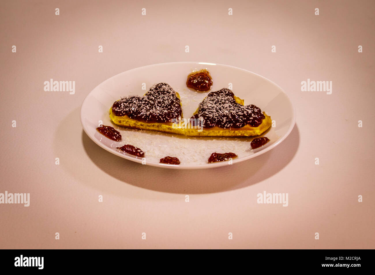 Due a forma di cuore fumetto waffels confettura di fragole e farina di cocco su di loro su sfondo bianco Foto Stock