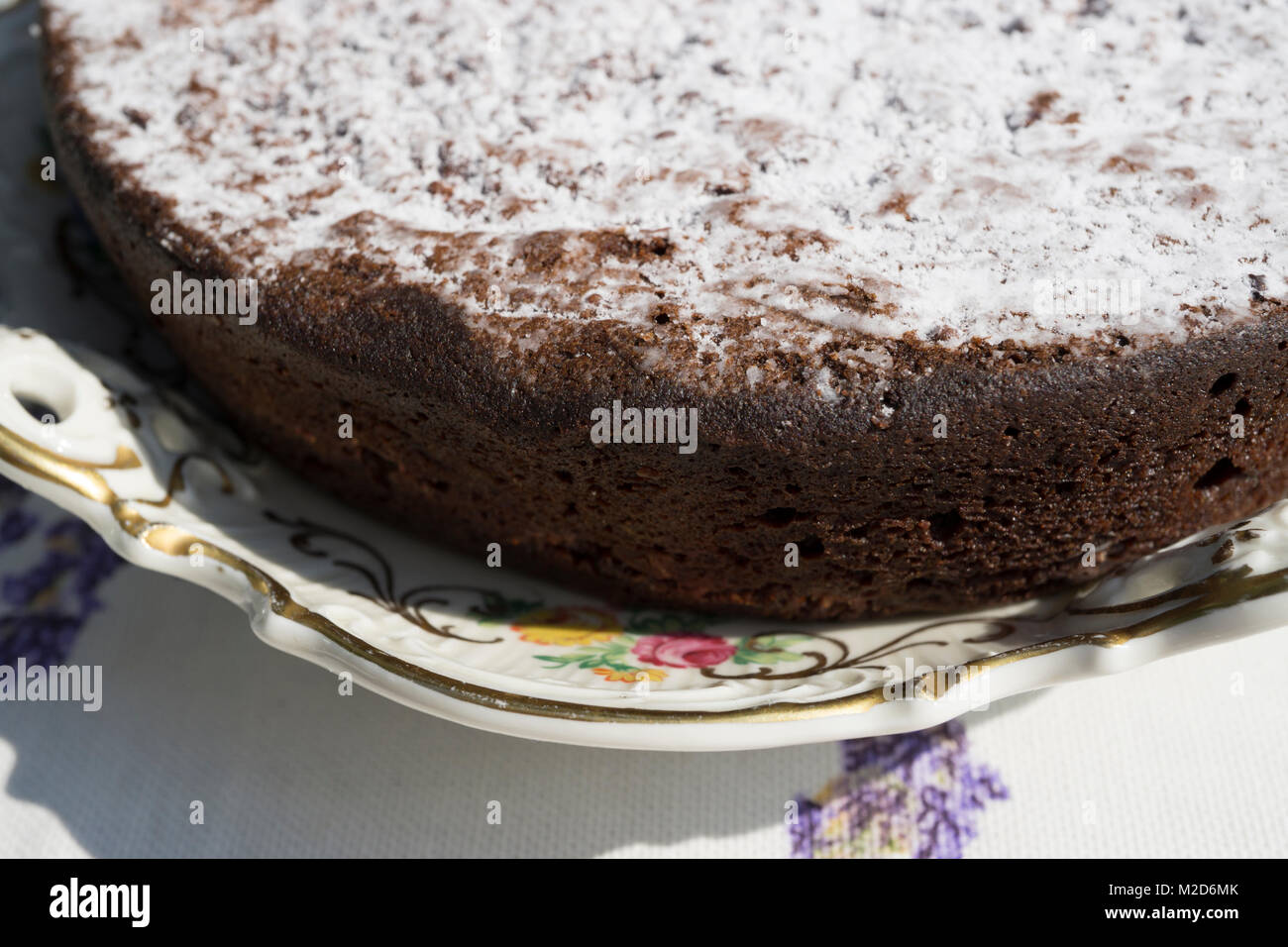 Primo piano di una torta al cioccolato spolverata con zucchero a velo su un antico piatto floreale. Si possono vedere bolle d'aria sul bordo della torta. Foto Stock