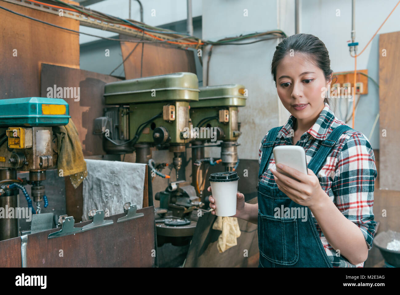 Sorridente bella macchina fresatrice di fabbrica lavoratore di sesso femminile il lavoro finito e tenendo calda tazza di caffè tramite telefono cellulare rilassante. Foto Stock