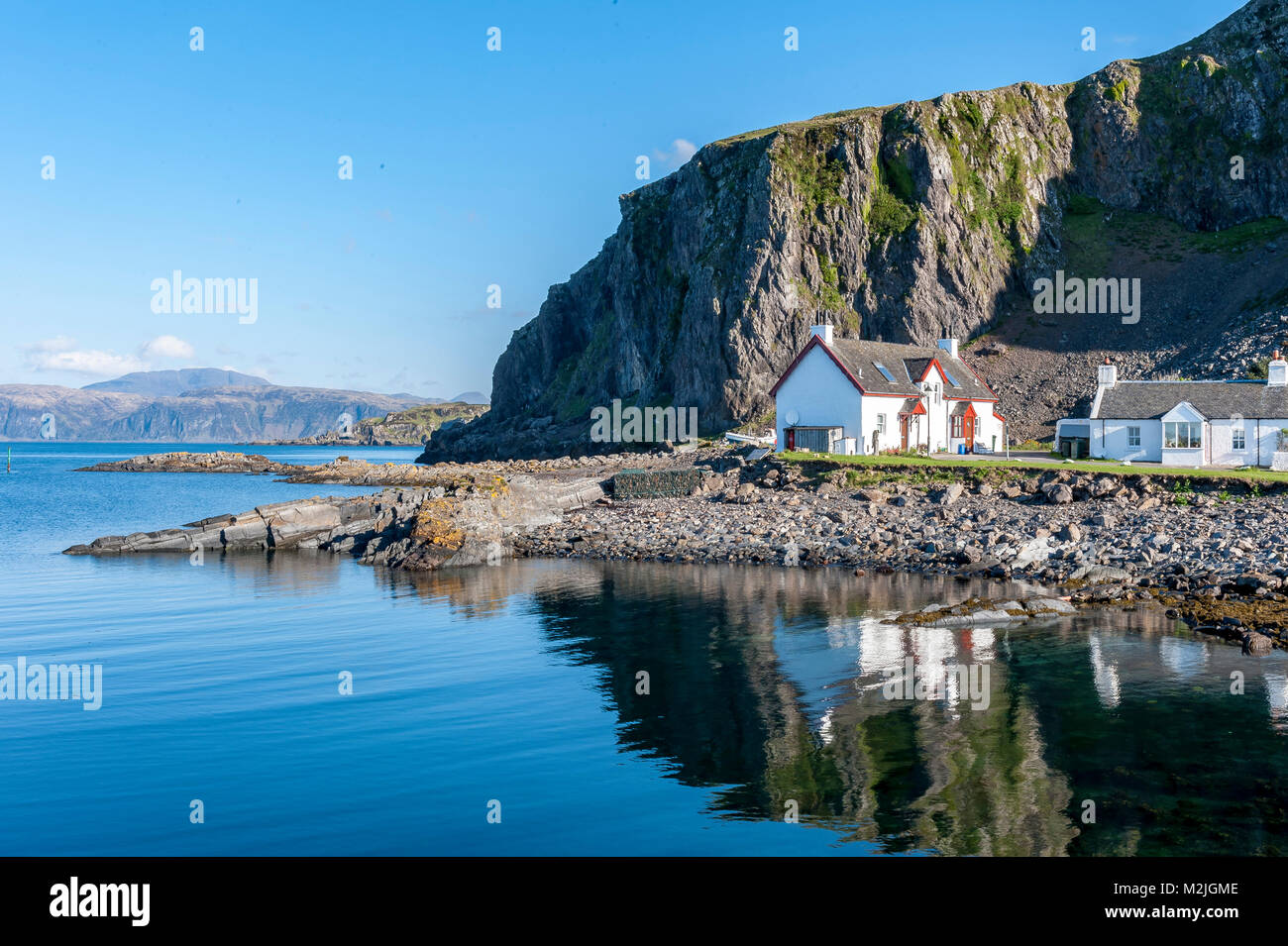 Splendida vista del villaggio di Ellenabeich Seil Isola, Scozia, con casette bianche alla base della scogliera di roccia si riflette nelle calme acque dell'oceano Foto Stock