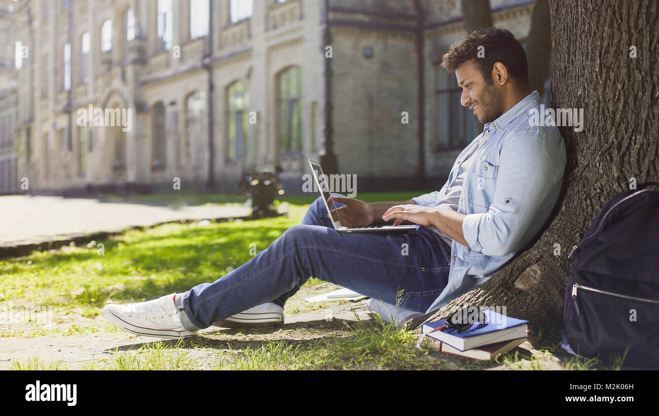 Giovani mixed-gara guy utilizzando laptop sotto l albero, sorridente alla buona novella, emozioni Foto Stock