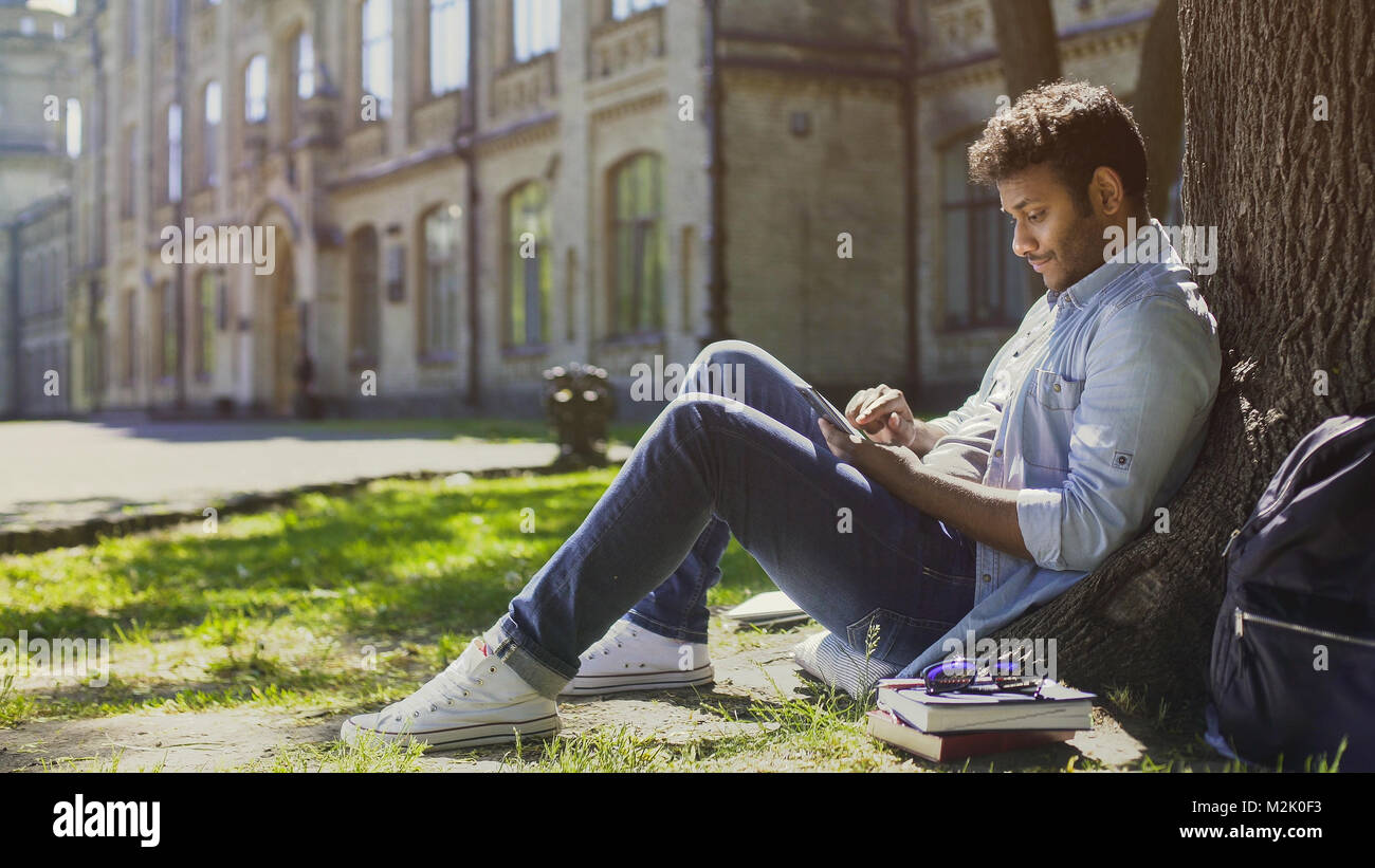 Giovane uomo seduto sotto agli alberi nel parco, utilizzando il telefono cellulare e sorridente, buone notizie Foto Stock