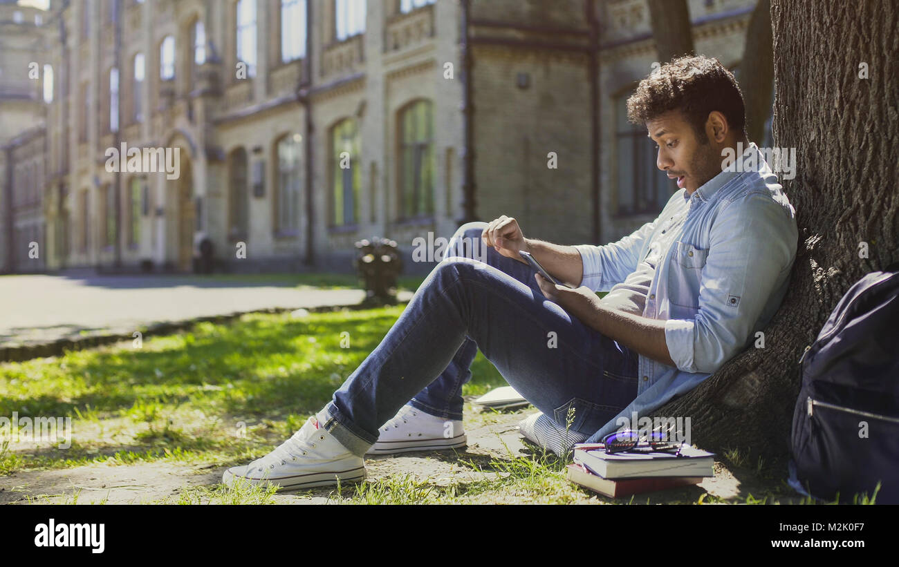 Ragazzo sorpreso seduto sotto agli alberi, utilizzando il telefono cellulare e il far reagire a newsfeed Foto Stock