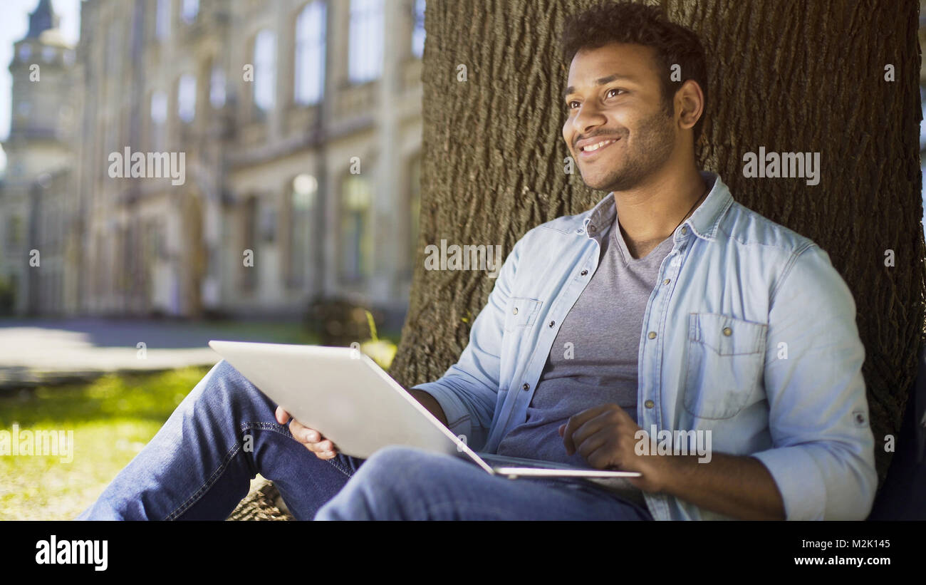 Mixed-gara giovane con laptop seduto sotto agli alberi, dating profilo sito Foto Stock