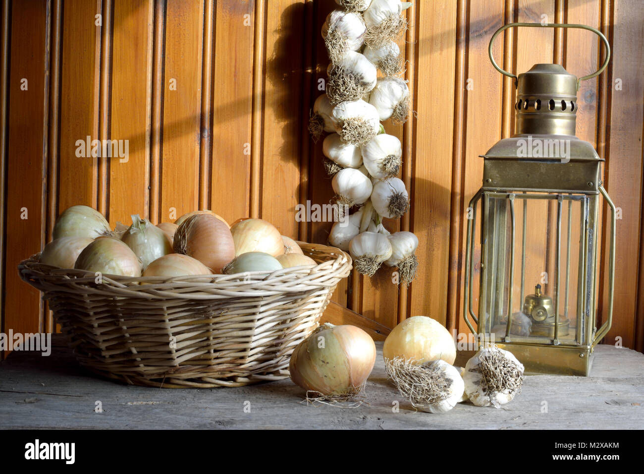 Le cipolle su un cestello, treccia di aglio appeso alla parete di legno e una lampada ad olio Foto Stock