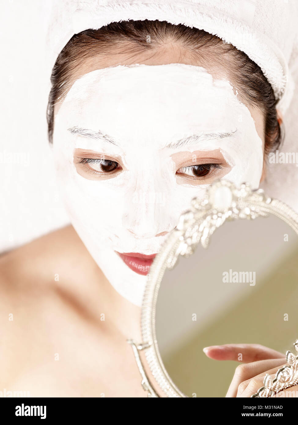 Bella giovane donna asiatica con maschera facciale giacente sul retro sul letto guardando nello specchio. Foto Stock