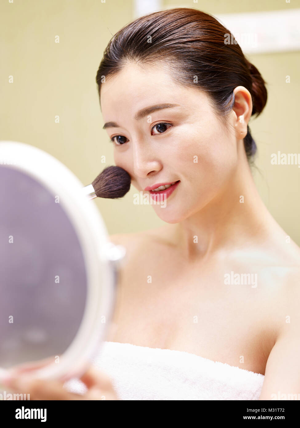 Bella giovane donna asiatica guardando nello specchio mentre si applica il make-up sul viso utilizzando una spazzola. Foto Stock