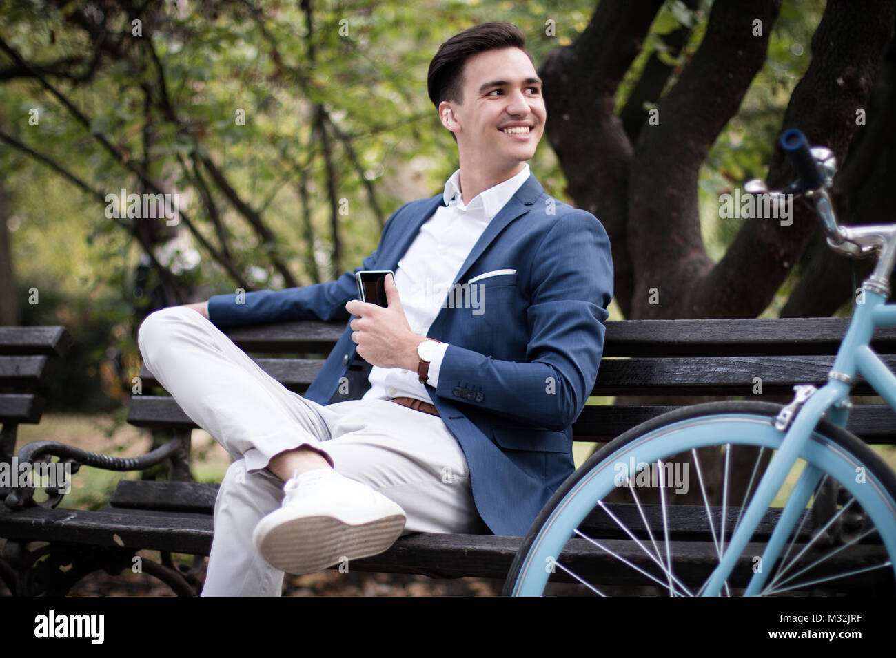 Giovane impreditore con lo smartphone seduta su una panchina in un parco con la sua bicicletta accanto a lui. Foto Stock