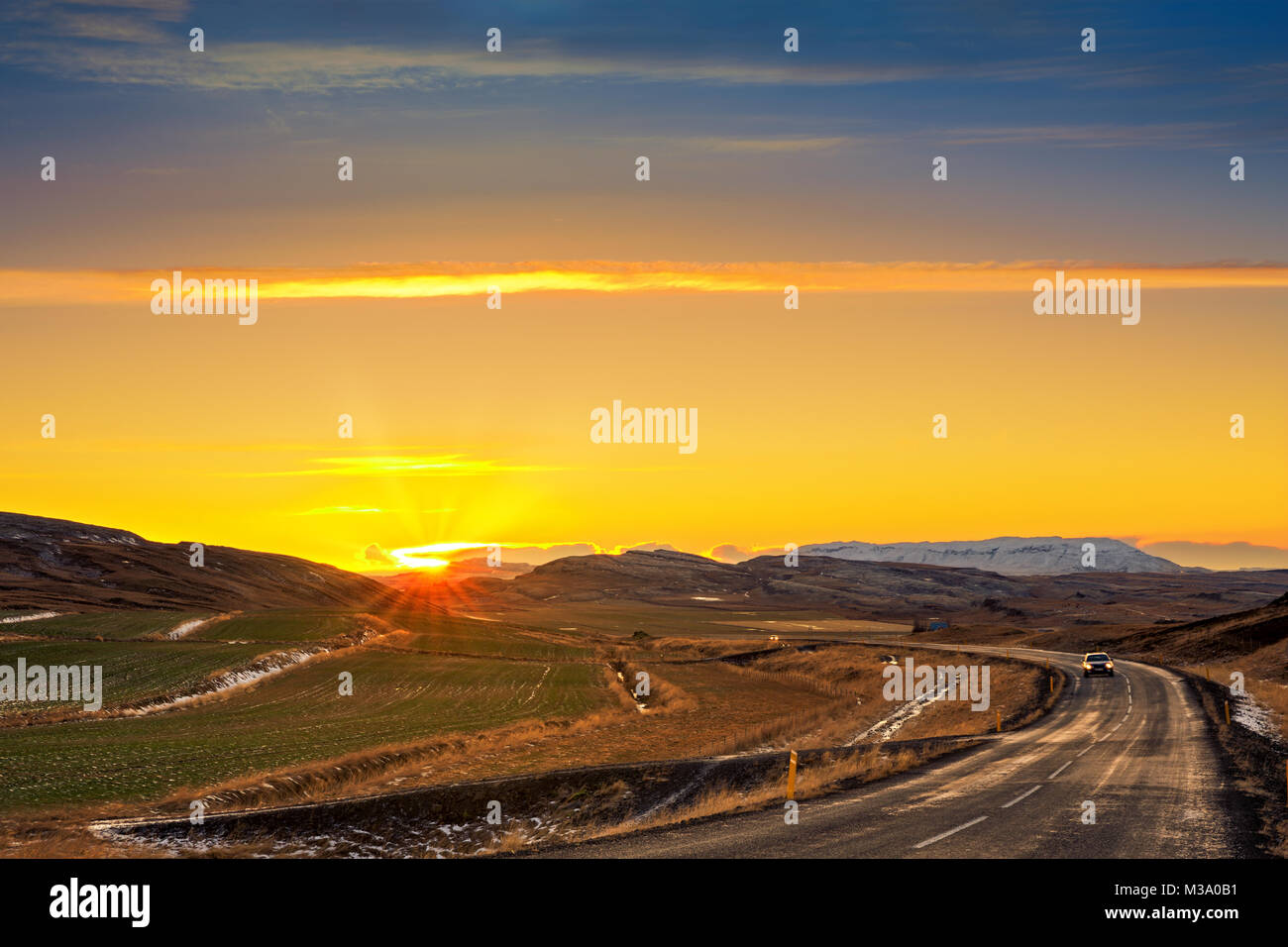 Strada tortuosa con lonely auto attraversa i campi agricoli e le montagne in Islanda rurale al tramonto. Foto Stock