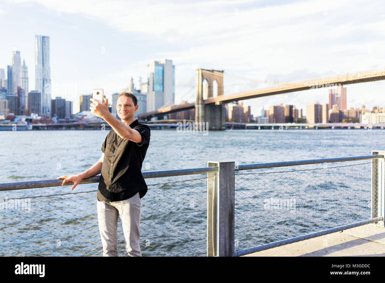 Giovane uomo fuori all'aperto in NYC New York City Ponte di Brooklyn Park da East River, ringhiera, guardando alla vista del paesaggio urbano skyline, tenendo selfie con Foto Stock