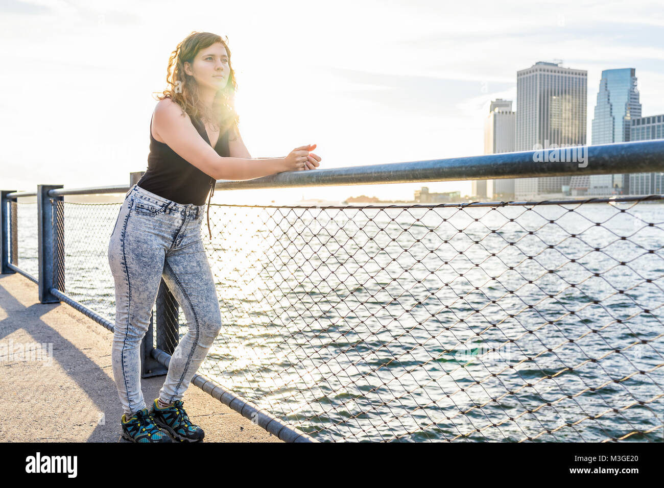 Giovane donna in piedi fuori all'aperto in NYC New York City Ponte di Brooklyn Park da East River, ringhiera, guardando alla vista del paesaggio urbano skyline, tramonto Foto Stock