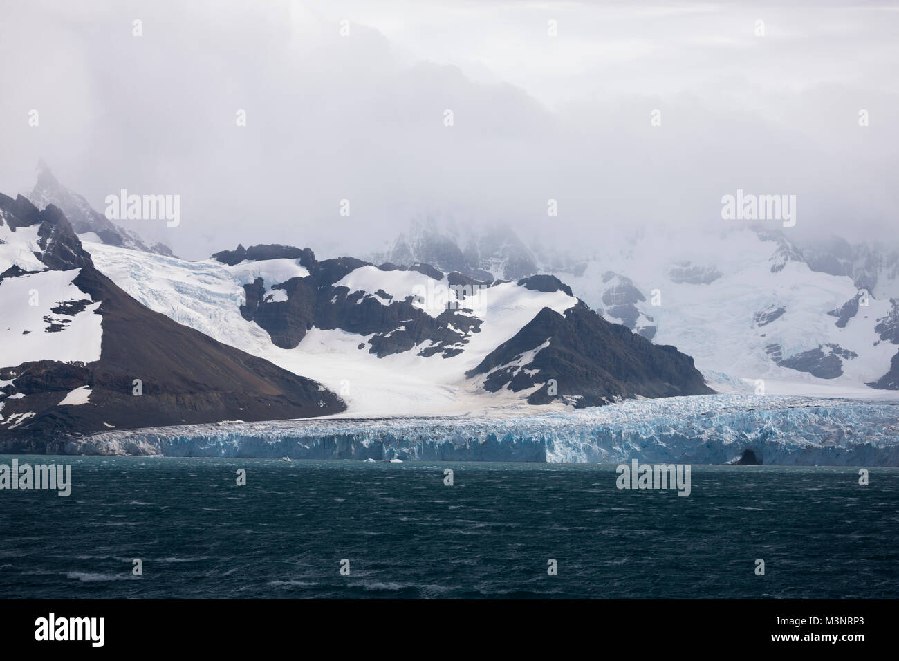 Big Blue Ice glacier contro le montagne nere grave tempesta di neve si avvicina oltre la montagna, oceano meridionale della Georgia del sud le isole, regioni antartiche Foto Stock