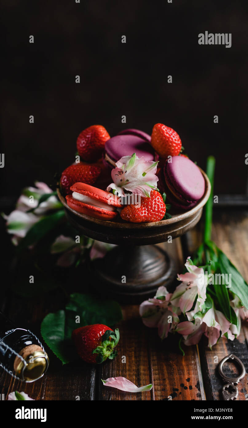 Ciotola con macarons e fragole su un tavolo di legno con fiori e bottiglia di champagne Foto Stock