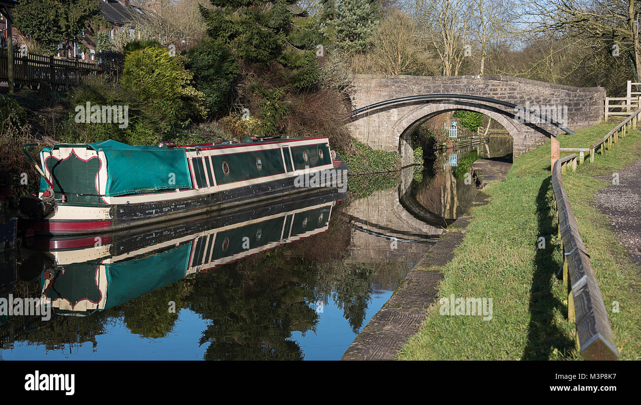 Una tranquilla Vista panoramica di un narrowboat con riflessioni ormeggiata su un canale con un ponte Foto Stock