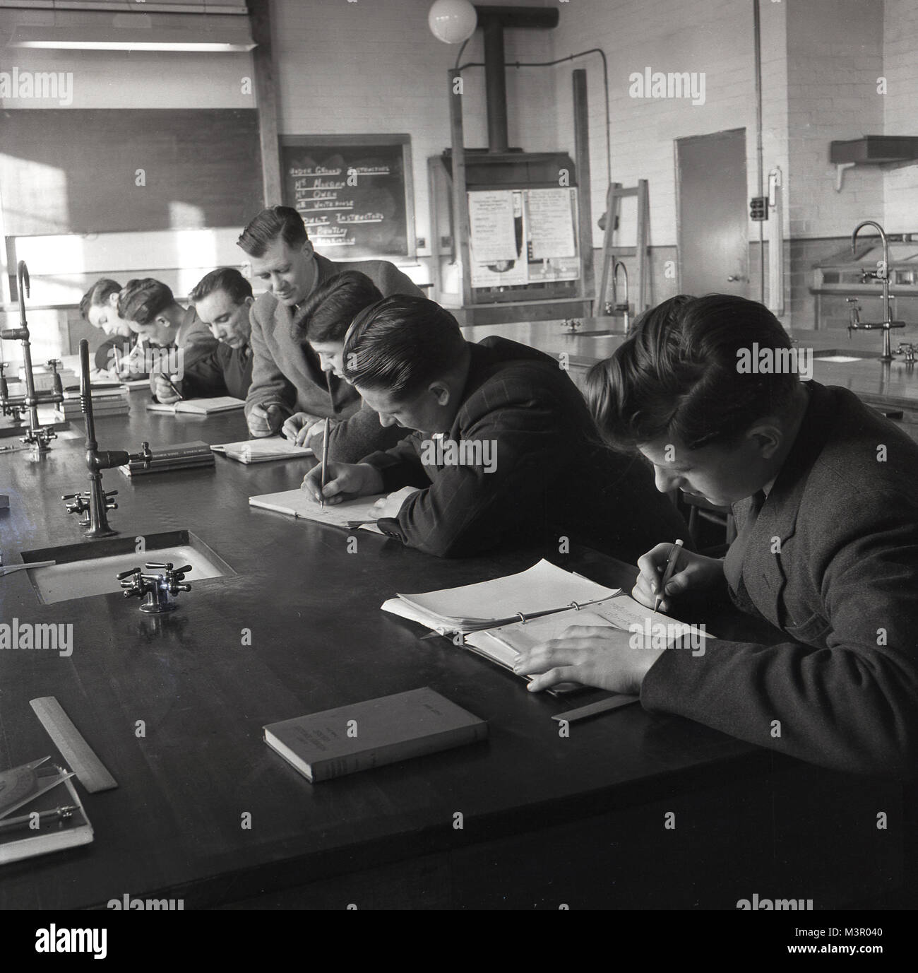 Degli anni Cinquanta, storico, giovane maschio scienza gli studenti seduti su un banco da lavoro in un laboratorio a un collegio tecnico scrivere note, Inghilterra, Regno Unito. Questi collegi offerto ulteriore istruzione per coloro che abbandonano modren secondario scuole. Foto Stock