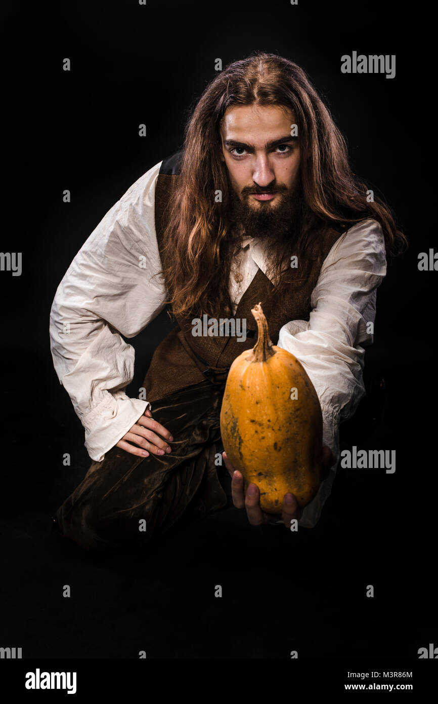 Ritratto di un uomo con la barba e capelli lunghi che indossa un medievale costume pirata su uno sfondo nero, un pirata tenendo una zucca matura Foto Stock